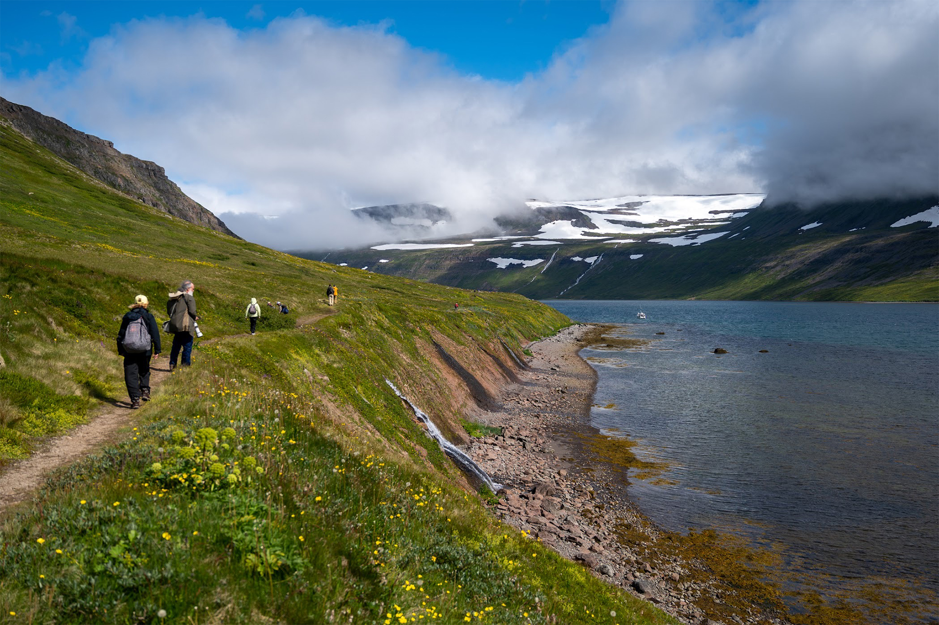 Group of travelers trek and hike on a single track in green meadows in iceland near river and flowing waterfalls